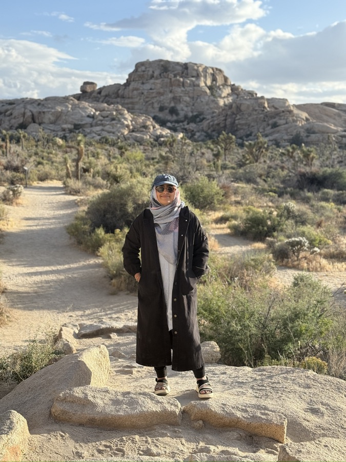 Mehvash standing on a boulder on a desert trail in Joshua Tree National Park, wearing a long dark coat and cap, with Joshua trees and scrub behind her.